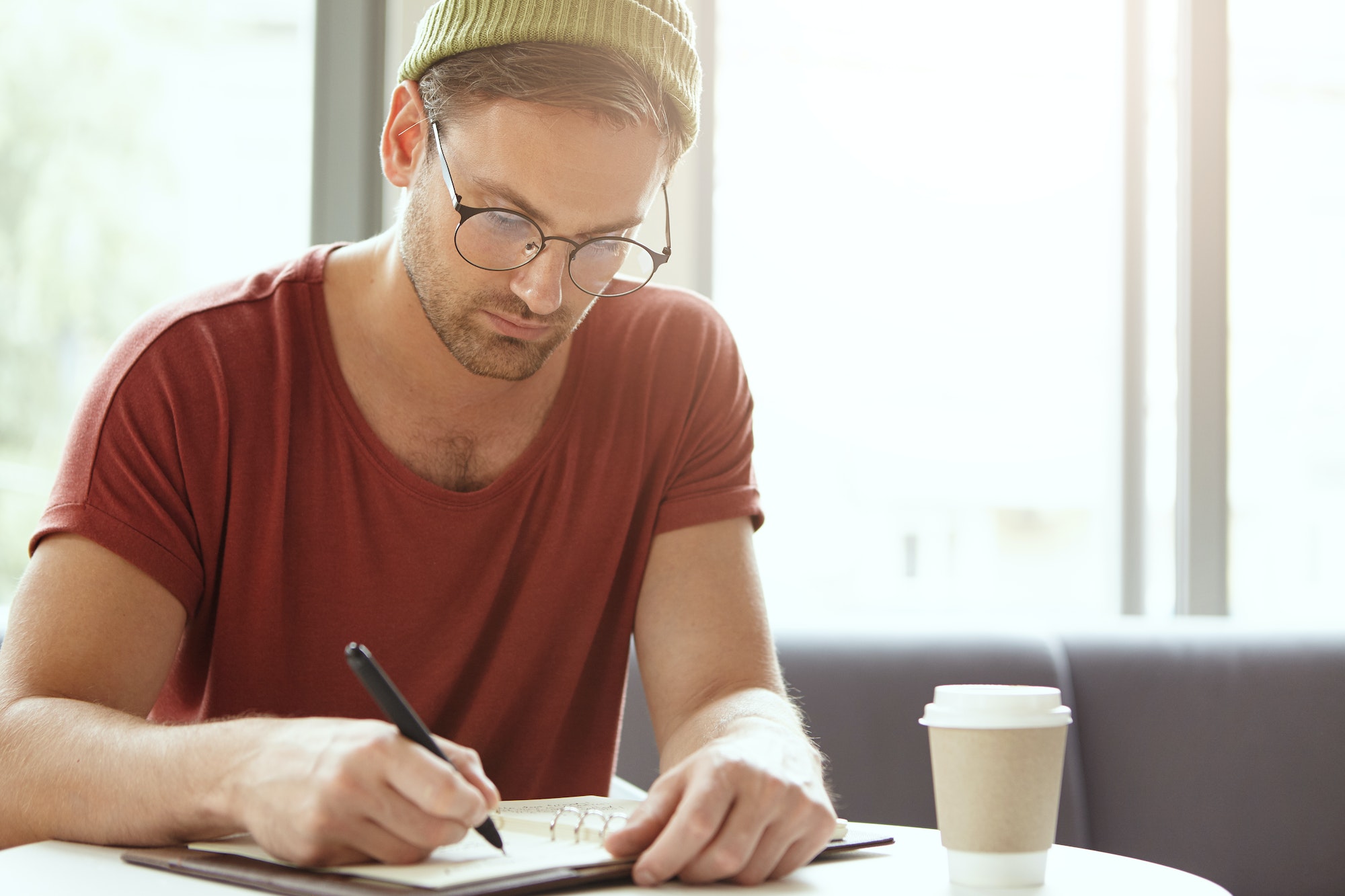 Young talented copywriter puts down ideas in notebook, sits at coffee shop, looks seriously at notes